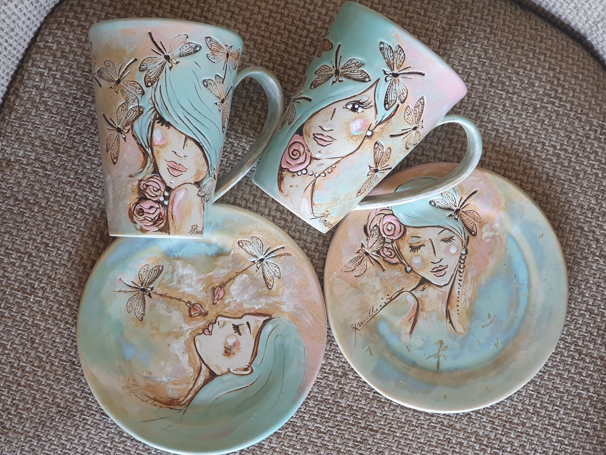 Ülaltvaade kahele keraamilisele tassile ja kahele taldrikule, mis asuvad tekstuuriga beežil kangal. Nõudel on pastelne piparmündiroheline ja roosakas glasuur. Tasse ja taldrikuid kaunistavad pruunide piirjoontega romantilised naisenäod, roosimotiivid ning detailsed kiilid.